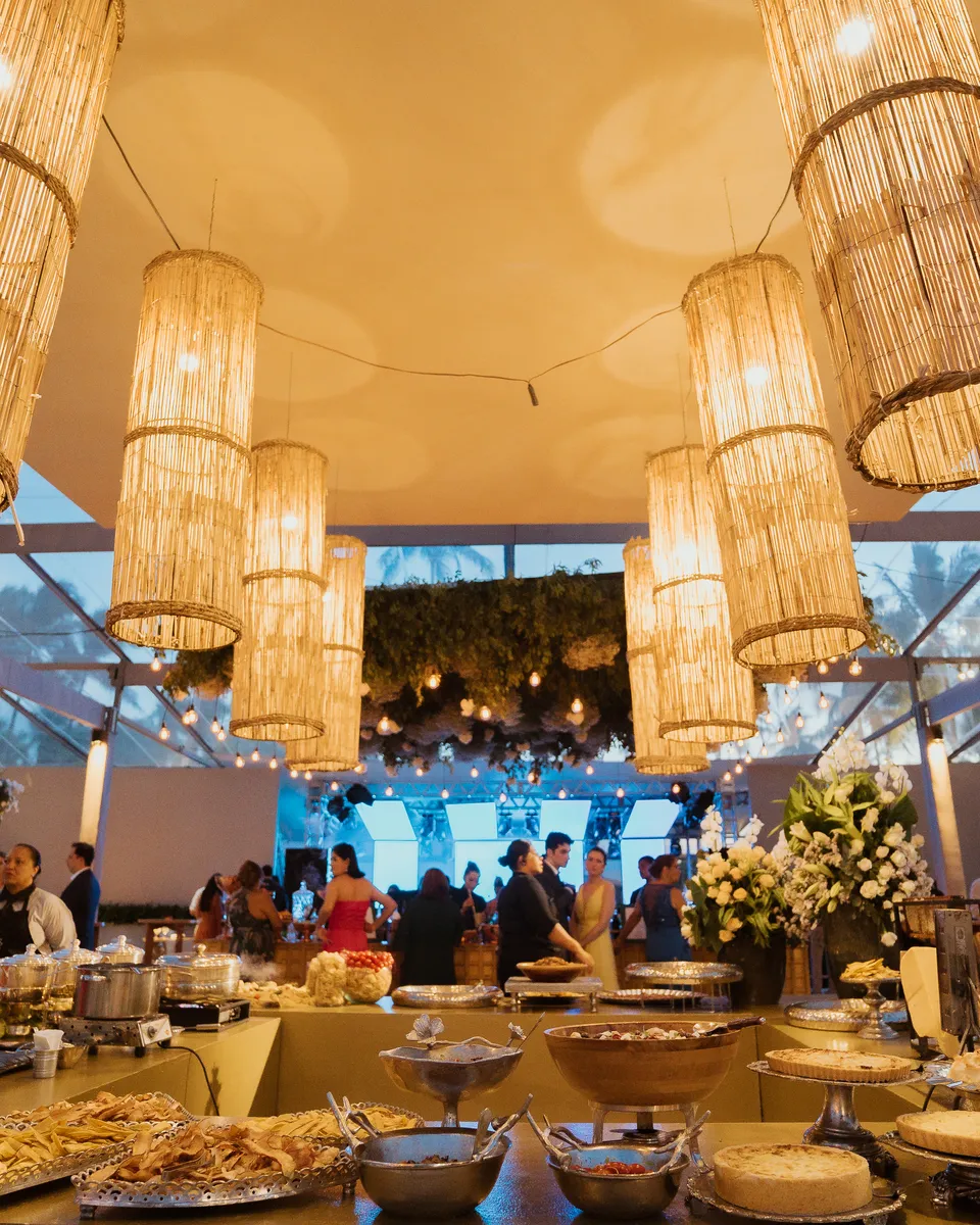 A tenda do casamento dos Medinas à noite, com luminárias de palha sobre a ilha do buffet servida e os convidados ao fundo