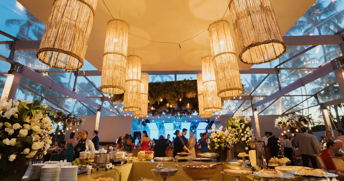 A tenda do casamento dos Medinas à noite, com luminárias de palha sobre a ilha do buffet servida e os convidados ao fundo
