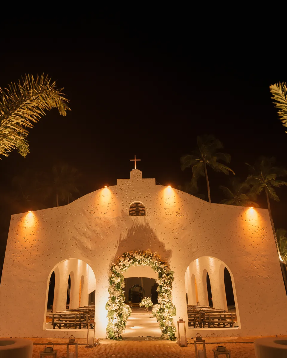 Capela do casamento dos Medinas iluminada à noite, entre coqueiros, na praia