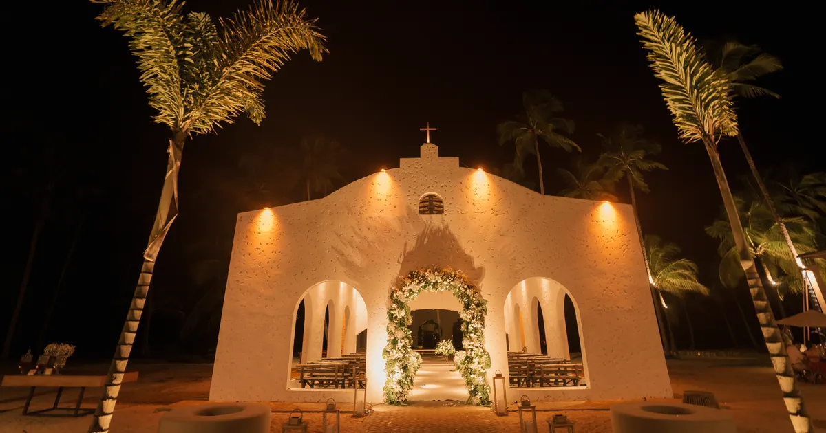 Capela do casamento dos Medinas iluminada à noite, entre coqueiros, na praia