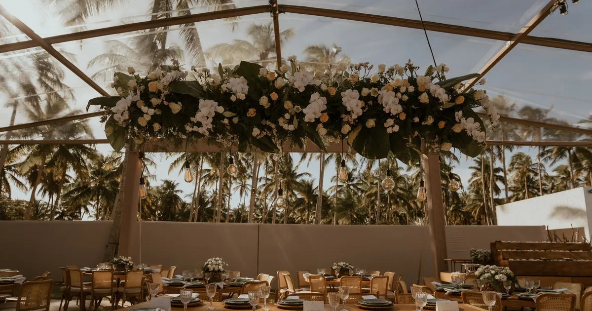Salão da recepção do casamento dos Medinas, com mesas postas, arranjo floral suspenso e coqueiros ao fundo